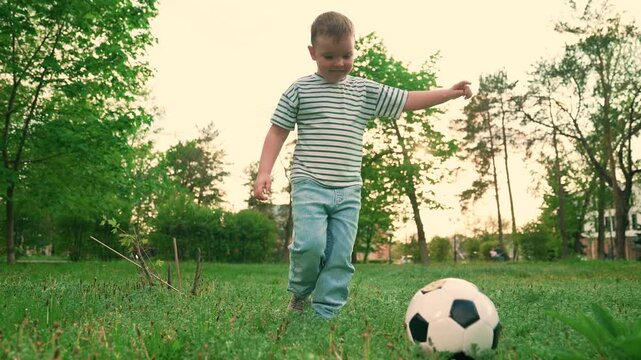 Little boy plays soccer on green grass. Kid plays football, city park. Child kicks soccer ball outdoors summer. Happy family. Athletic child leads active lifestyle. Kid boy footballer city park, sport
