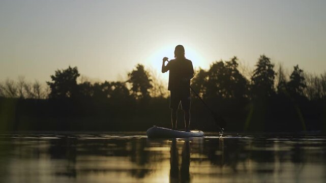Supboard boad man. A man paddles on a calm lake as the sun sets behind him. The man enjoys a serene moment with nature, surrounded by trees and a peaceful atmosphere.