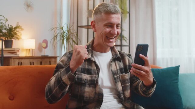 Surprised excited young man winner holding smartphone reading good news amazed by online bet bid game win. Happy guy male on sofa looking at screen overjoyed by victory success in home apartment.