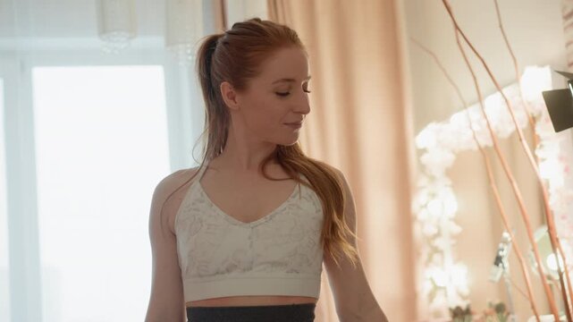 Woman in sports bra performs bicep curl exercise indoors. Bright home environment features natural and artificial lights curtains large window decor elements like branches with flowers