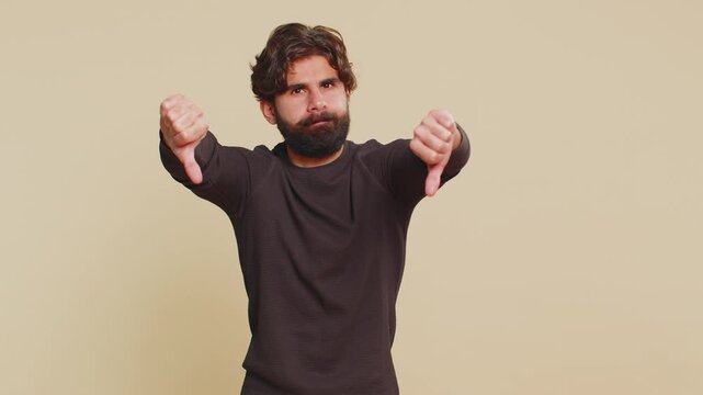 Dislike. Upset unhappy Lebanese man in long sleeves showing thumbs down sign gesture, expressing discontent, disapproval, dissatisfied, dislike. Handsome young guy. Indoors studio on beige background.
