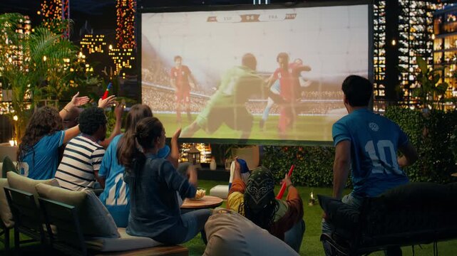 Soccer supporters celebrate favorite team scoring goal during match projected at rooftop watch party; elated spectators jump and hug, feeling ecstatic during live soccer game broadcast.