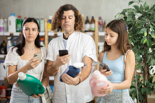 Friendly modern family of three using smartphones to scan barcode on cleaning supplies bottles in local supermarket, looking for discounts, checking for digital coupons and special offers