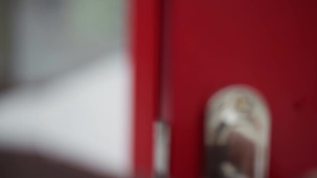 Close up shows hands reaching for and grasping shiny metal handle on bright red wooden door gradually pulling it to open. focus remains tightly on interaction between hand and hardware