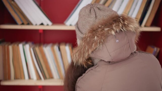 Shows part of young woman head and upper body as woman faces away from camera wearing winter clothing with bookshelves in background back coat hat bookshelf