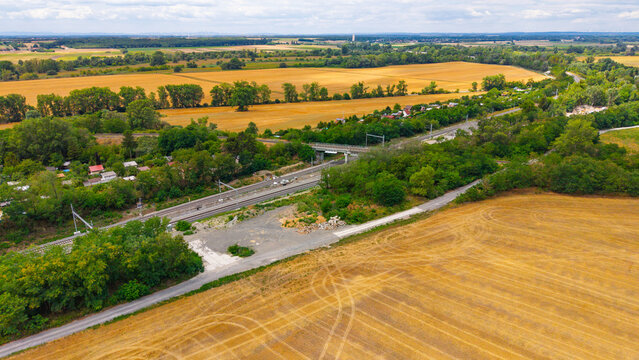 Aerial view of railway line passing through countryside. Train tracks cut across green trees between harvested wheat fields under a cloudy sky.
