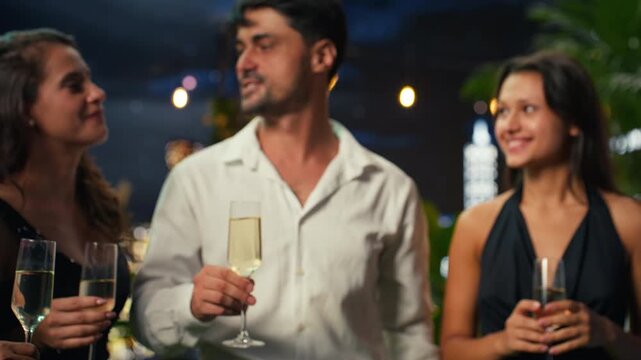 Close up of glasses of champagne clinked by friends celebrating anniversary on rooftop terrace at night, with party guests toasting with sparkling wine to mark important milestone.