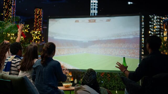 Football supporters react with disappointment after conceding a goal while watching a projected match together on a rooftop terrace above the city.
