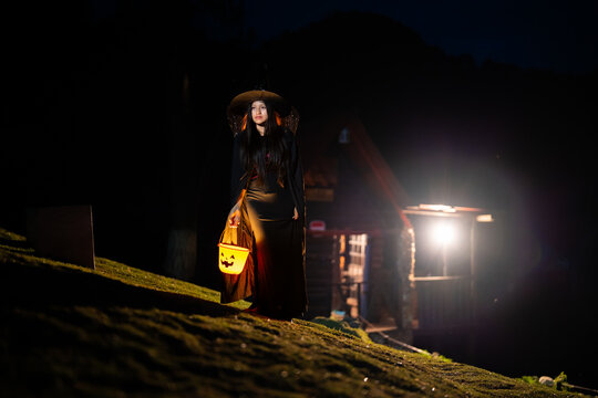 Witch walking on a hill at night with a lit-up pumpkin bucket and a rustic cabin in the background on Halloween.