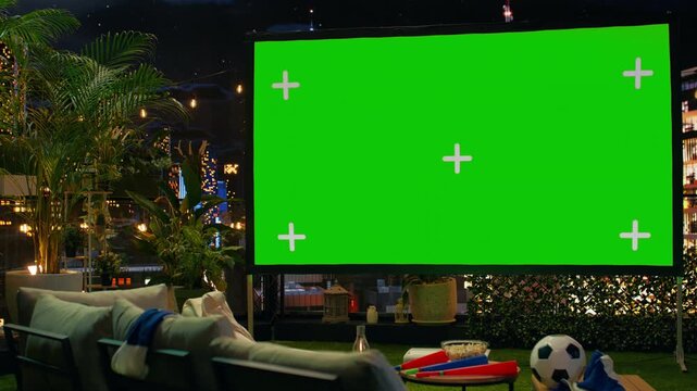 Blank projection screen mockup in rooftop lounge next to fan props prepared for football match viewing; chroma key display provides space for soccer match broadcast during watch party.