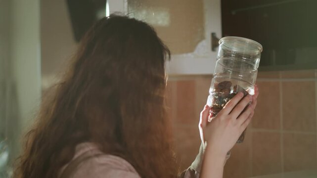 Woman examines content of large clear glass jar filled with what appears to be preserved or dehydrated foods. Woman holds it carefully under soft natural light in indoor kitchen setting