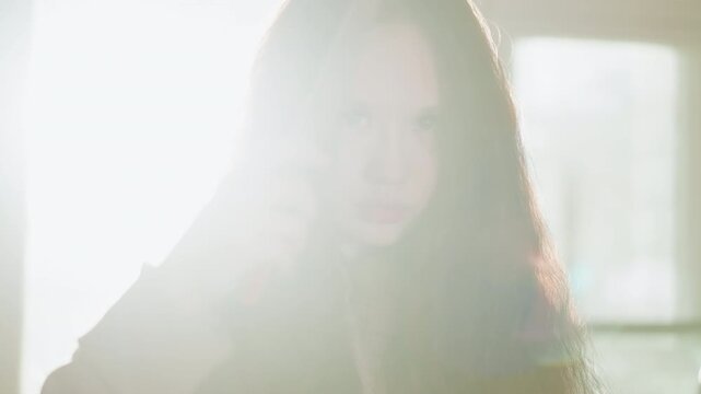Woman holds knife indoors under bright light. Close up shows her focused eyes behind long hair as woman grips blade. Soft sunlight streams through window in background creating dramatic
