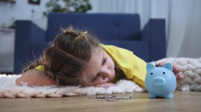Child saving coins in piggy bank at home. Girl puts coin into blue piggy bank. Kid learns saving money. Little girl lies on floor counting coins. Child collects money for future savings.