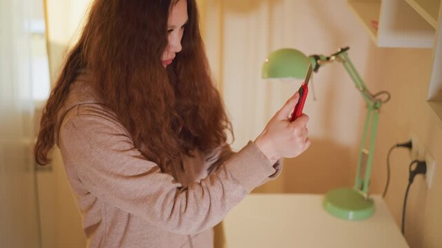 Woman stands at white table examining or preparing to use small red utility knife. Art supplies like pencils paint containers and paper are visible on tabletop under warm lighting