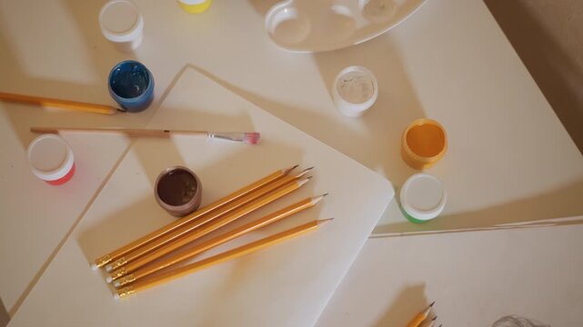 Pencils are arranged near drawing paper with incomplete pencil portrait. Art tools like paints and brush are visible in studio setting showing art creation materials on table
