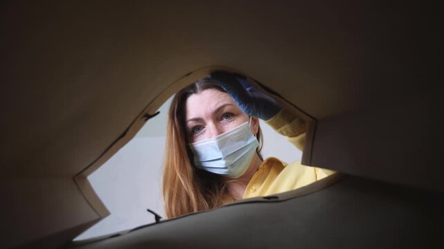 Woman in mask opens cardboard box. Gloved hand reaches into parcel. Masked woman looks inside package during safe delivery. Woman unpacks box wearing protective glove. Unboxing parcel pov view.