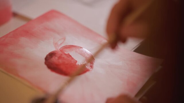 Close up shows someone using paint to create image focusing on process and materials involved in art making. Hands hold tools like brushes while working on canvas or paper with visible