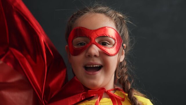 Young girl wearing bright red superhero mask and flowing cape with yellow outfit, posing confidently against dark background. Smiling child embodies hero character in imaginative play concept.