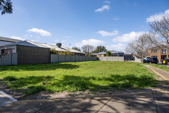 An vacant lot between houses and boundary fences along a quiet suburban street, with established homes and parked cars nearby. An undeveloped infill block within a maturing residential neighbourhood