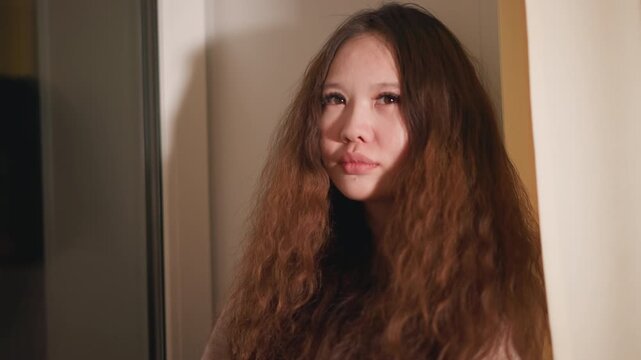 Close up shows upper body and face area of young woman in indoor setting. Her expression is neutral to slightly pensive as woman stands near doorway with soft lighting creating gentle