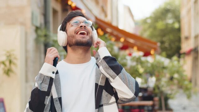 Happy overjoyed Indian man wearing wireless headphones choosing, listening energetic disco rock n roll music dancing outdoors. Hispanic young guy tourist standing in urban city street. Lifestyles.