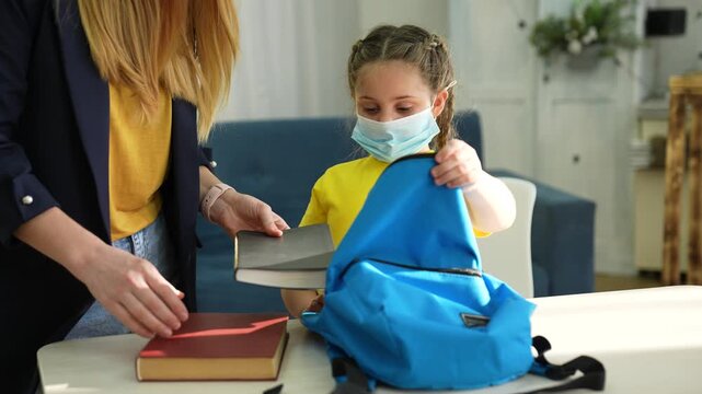 Mother helps daughter pack school backpack with books. Girl wearing face mask holds blue backpack open. Mom places books into bag. Parent preparing child for school during pandemic at home.