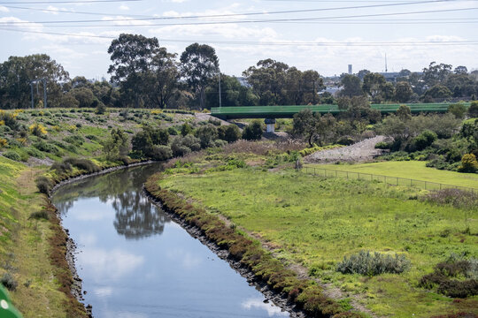 The curving channel of Stony Creek in Yarraville, Melbourne, Victoria,&nbsp;Australia&nbsp;surrounded by revegetated grassy banks, native trees,&nbsp;and a pedestrian bridge crossing in the background.
