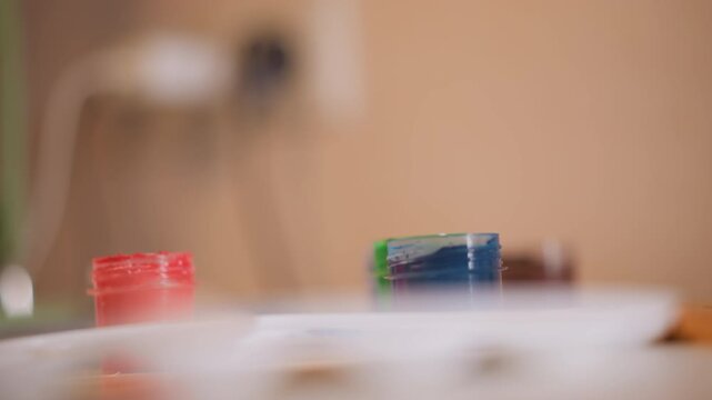 Close up focus remains steady across small containers of orange red blue green art paints arranged in soft focus sequence. Background stays blurred with neutral tones emphasizing still life