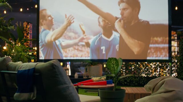 Football game broadcast is projected in an empty rooftop lounge at night, with comfortable seating and fan props arranged for a cozy soccer watch party.
