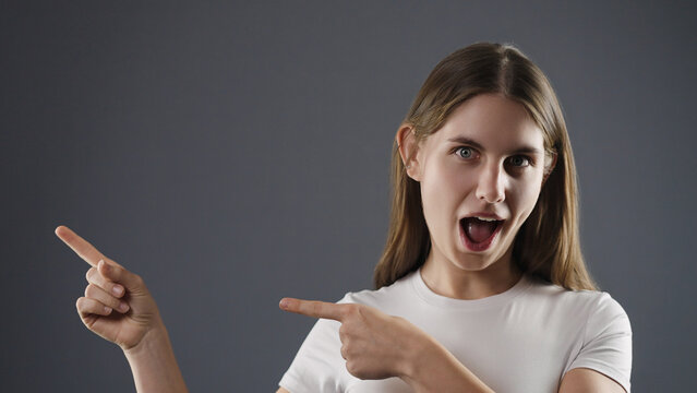 Young woman pointing fingers to the left with an open mouth and wide eyes on a gray background