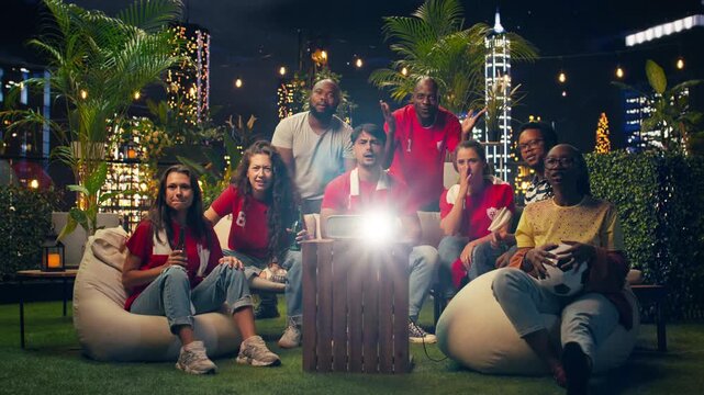 Happy football supporters cheer enthusiastically around projector during rooftop soccer match; joyful supporters celebrate team goal while enjoying evening watch party above city at night.