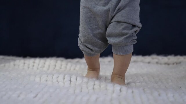 Baby feet walking barefoot on soft blanket. Toddler takes first steps on white knitted rug. Small bare feet step on cozy wool surface. Infant feet and tiny toes visible. Baby walks indoors at home.