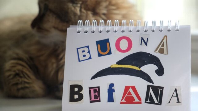 Cozy home moment: cute cat napping beside a child&rsquo;s handmade &ldquo;Buona Befana&rdquo; greeting. The letters are cut from magazines and carefully glued onto the paper, giving the note a tender, handmade charm.