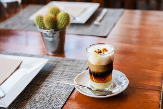 A barraquito coffee stands on a wooden restaurant table with its layers of milk coffee and foam clearly separated. Local drinking culture for travel dining and island holiday content.