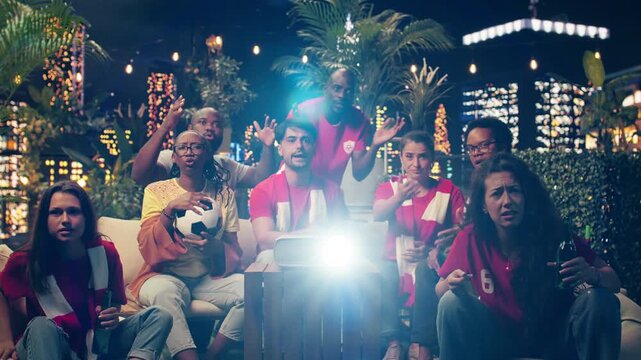Concerned football supporters react tensely during a rooftop soccer broadcast after a defensive blunder, watching the live match projection under the night sky.