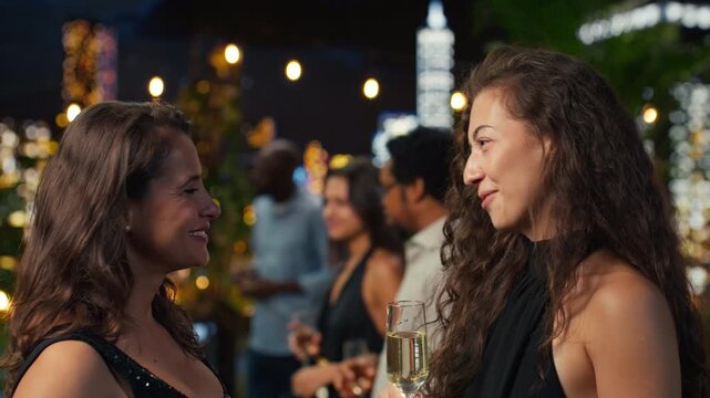 Close up of women sharing stories over champagne at a rooftop party in a modern terrace lounge above the illuminated city skyline at night.