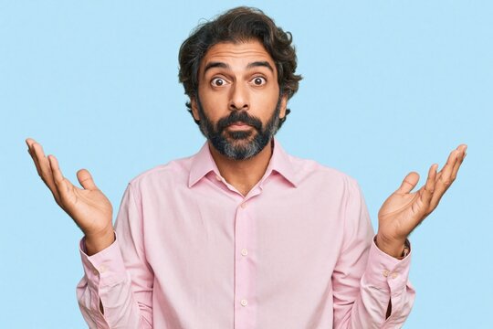 Handsome middle age man with beard wears pink shirt. Shows confused expression, shrugs shoulders against blue background. Puzzled male makes clueless gesture with raised hands. Uncertain person feels