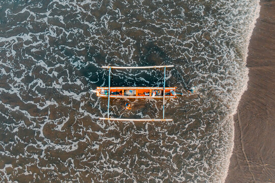 Direct overhead drone view of traditional orange jukung outrigger boat floating in foamy sea water near black sand beach. Fishermen pushing boat into ocean waves in Bali