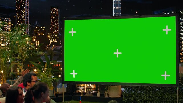 Green screen projection on rooftop terrace used by group of friends enjoying soccer match together; supporters looking at live football game broadcast on mockup display above the city skyline.