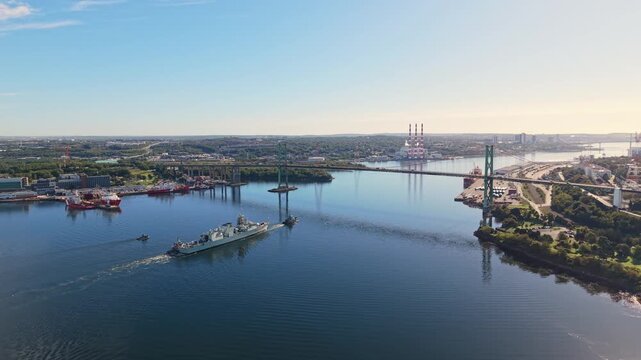 Aerial view of a military towing operation in Halifax Harbour, Nova Scotia, with a military ship pulling another vessel beneath a bridge while creating long wakes across the calm morning water.