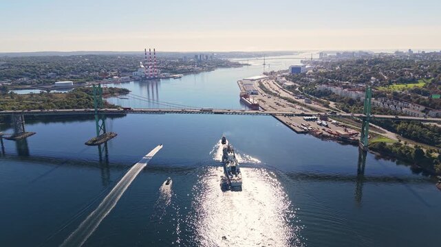 Aerial cinematic footage of a military ship and towed vessel traveling together through Halifax Harbour, Nova Scotia, passing beneath a bridge in the soft light of early morning.