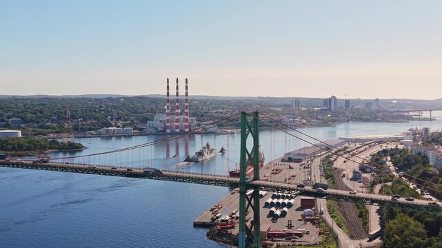Drone view of a military ship towing a large vessel beneath a bridge in Halifax Harbour, Canada, with calm morning water, soft light, and the Halifax waterfront visible in the distance.