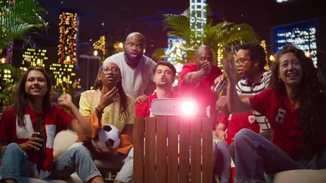 Joyful soccer supporters watch a projected match and cheer together as friends clap after their favorite team scores a goal during the celebration.