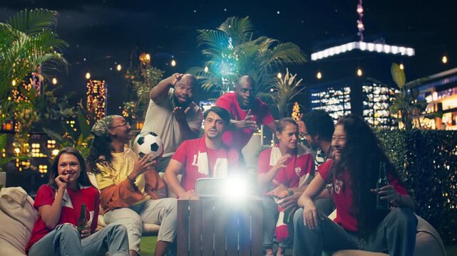 Excited football supporters follow soccer action while standing around rooftop projector, enjoying goal scored during tournament game and drinking beer at an evening rooftop watch party.