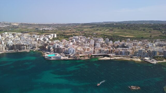 St Paul's Bay and Bugibba, Malta waterfront with apartment buildings, marina and boats along the turquoise Mediterranean coast on a sunny day, tracked by drone.