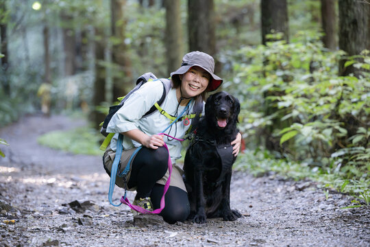 愛犬の大型犬と登山を楽しむ飼い主の全身カメラ目線で座る、森林の中アウトドアやトレッキングのレジャーイメージ
