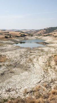 Low water level exposes cracked shoreline around shrinking reservoir in dry hilly landscape. Drought water scarcity, climate risk, environmental crisis drive conservation awareness resource management
