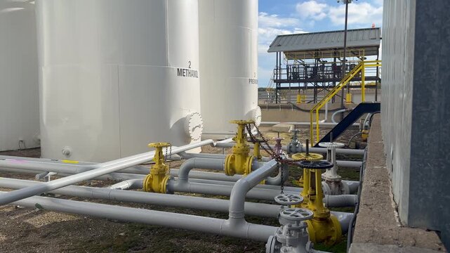 A pan left with a slow push in on an industrial methanol storage facility, showcasing the tanks and piping systems under a clear blue sky.