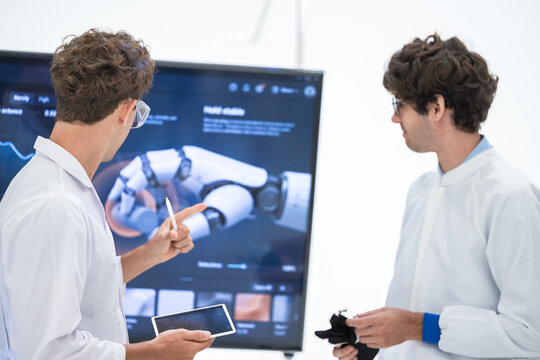 Two young engineers compare tablet notes and a black robotic glove in front of a large robotic hand render on screen in a bright robotics lab, a next generation prosthetics concept