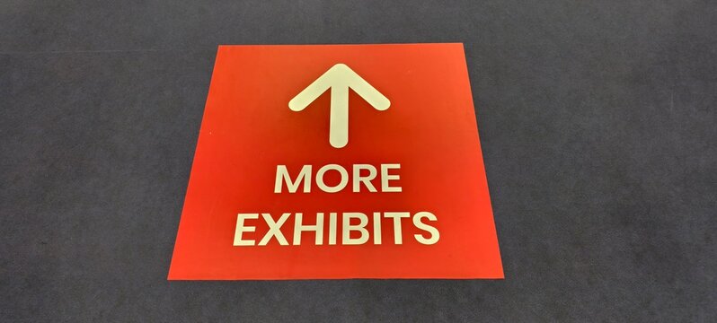 Red rectangular floor sign with bold white text and upward arrow reading &ldquo;MORE EXHIBITS.&rdquo; Designed for clear navigation in museums, galleries, or exhibition spaces.